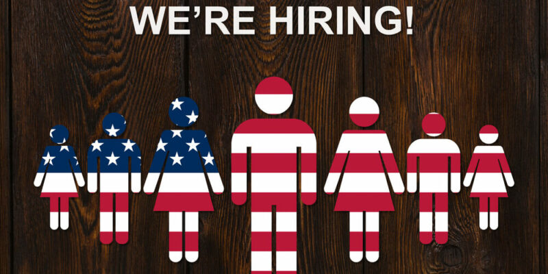 Paper men colorized into the color of the usa flag on wooden background with text WE'RE HIRING. Business concept.