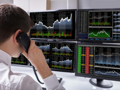 Stock Broker Talking On Telephone In Front Of Multiple Computer Screen Showing Graphs