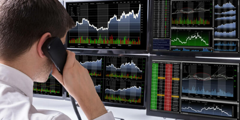 Stock Broker Talking On Telephone In Front Of Multiple Computer Screen Showing Graphs