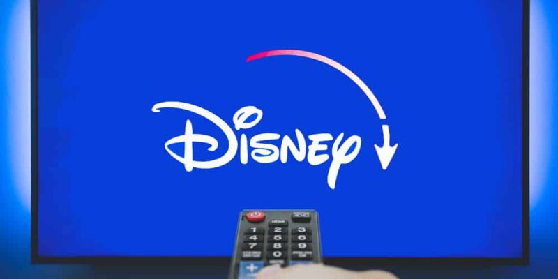 Man holds a remote control. Disney+ logo on TV screen in background.